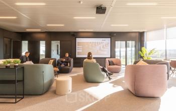 Lichte, moderne kantoorruimte te huur op Korte Keppestraat 19 in Aalst, met comfortabele zitplaatsen en een groot presentatiescherm. Drie personen zijn in gesprek en voeren zakelijke activiteiten uit.
