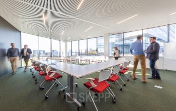 Ruime vergaderruimte op Korte Keppestraat 19 in Aalst, met glazen wanden en rode stoelen, waar meerdere mensen zakelijke activiteiten uitvoeren. Kantoorruimte te huur met moderne faciliteiten.