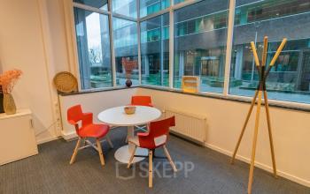 In deze kantoorruimte te huur op Uitbreidingstraat 84 in Antwerpen Berchem, zie je een kleine conferentiehoek met een ronde tafel, vier rode stoelen en een kapstok. Grote ramen zorgen voor veel natuurlijk licht.