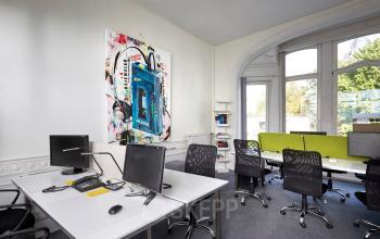 Kantoorruimte te huur in Brussel Etterbeek, Priester Cuypersstraat / Rue Abbé Cuypers 3. Moderne werkplek met bureaus, stoelen en computers, verfraaid met kleurrijke kunstwerken en grote ramen.
