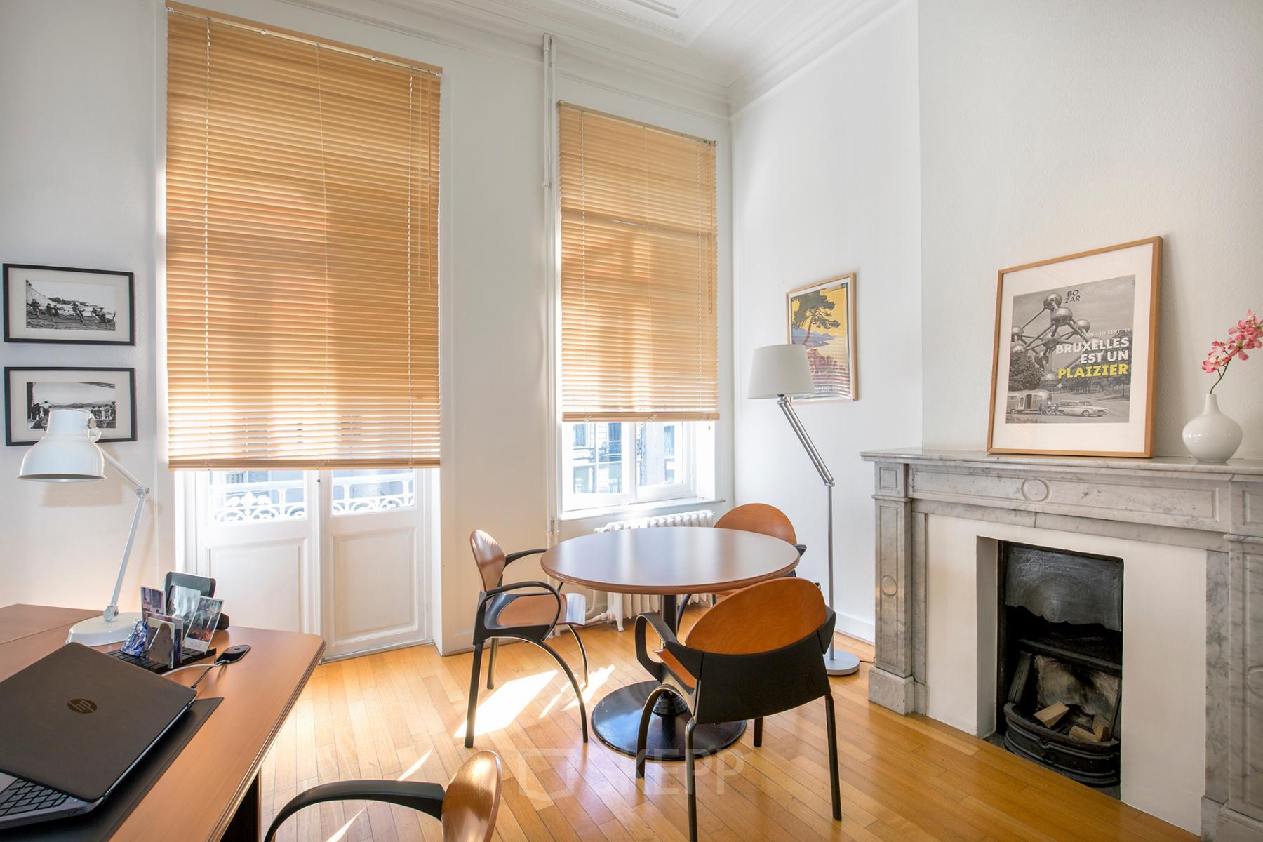 Kantoorruimte te huur in Brussel Europese Wijk, Breydelstraat / Rue Breydel 34-36, lichte kamer met houten vloeren, grote ramen met jaloezieën, ronde tafel met stoelen, bureau, en decoratieve open haard.
