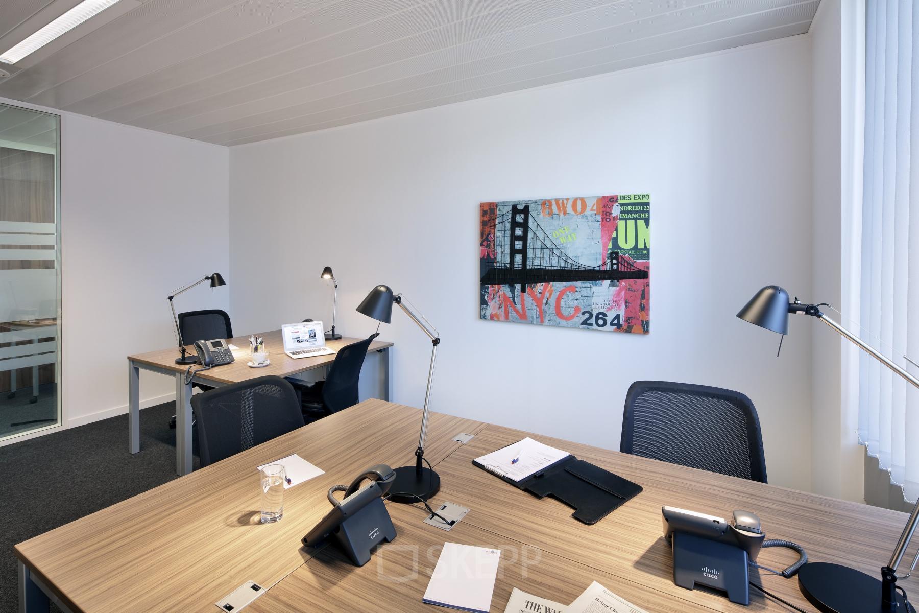 Moderne gemeubileerde kantoorruimte te huur met vier werkplekken en kleurrijk schilderij aan de muur, gelegen op Place Marcel Broodthaers 8 in Brussel-Zuid.