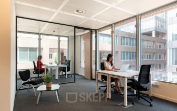 Kantoorruimte te huur op Gaston Crommenlaan 8, Gent Ledeberg. Moderne werkruimte met grote ramen. Twee medewerkers in overleg in de vergaderruimte, en één persoon aan het werk aan haar bureau.