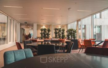 Kantoorruimte te huur aan Oktrooiplein 1 in Gent, licht en ruim met moderne meubels en grote ramen. Mensen overleggen aan een tafel bij de planten. Verrassend interieur voor zakelijke doelen.