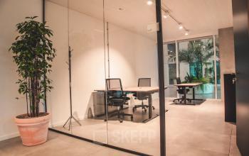 Kantoorruimte te huur in Gent, gelegen op Oktrooiplein 1. Moderne ruimte met glazen wanden, twee bureaus en stoelen, een grote plant en helder verlicht door plafondspots.