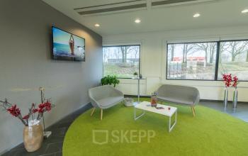 Lounge-ruimte in een kantoor te huur op Atealaan 34A, Herentals met moderne stoelen, salontafels, een groene tapijt en een tv aan de muur.